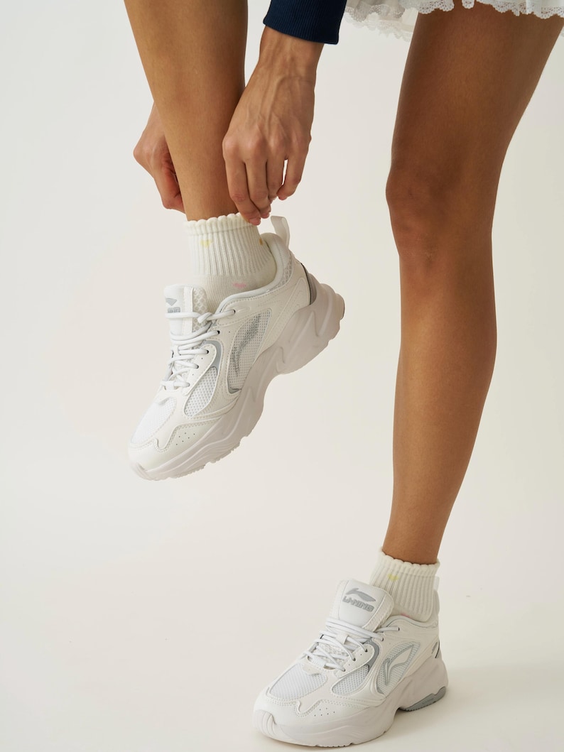 May include: White athletic sneakers with a chunky sole and mesh detailing. The sneakers have a lace-up closure and are paired with white ribbed socks. The shoes are a modern style, suitable for athletic activities or casual wear.