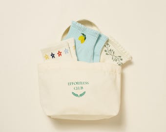 May include: A cream-colored tote bag with the text "EFFORTLESS CLUB" in green, holding four pairs of socks. The socks feature star, lemon, and floral designs in various colors. The bag has a simple, minimalist design.