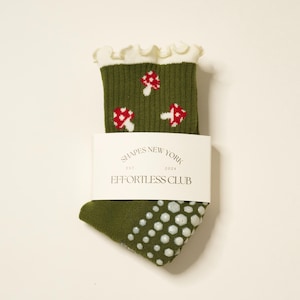 May include: Olive green socks with a white ruffled cuff and a pattern of red and white mushrooms. The socks have white non-slip grips on the sole and a white label that reads "SHAPES NEW YORK EST. 2024 EFFORTLESS CLUB."