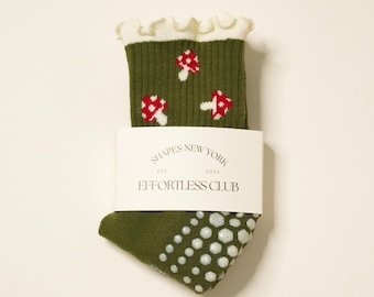 Puede incluir: Calcetines verde oliva con un puño blanco con volantes y un estampado de setas rojas y blancas. Los calcetines tienen agarres antideslizantes blancos en la suela y una etiqueta blanca que dice "SHAPES NEW YORK EST. 2024 EFFORTLESS CLUB."