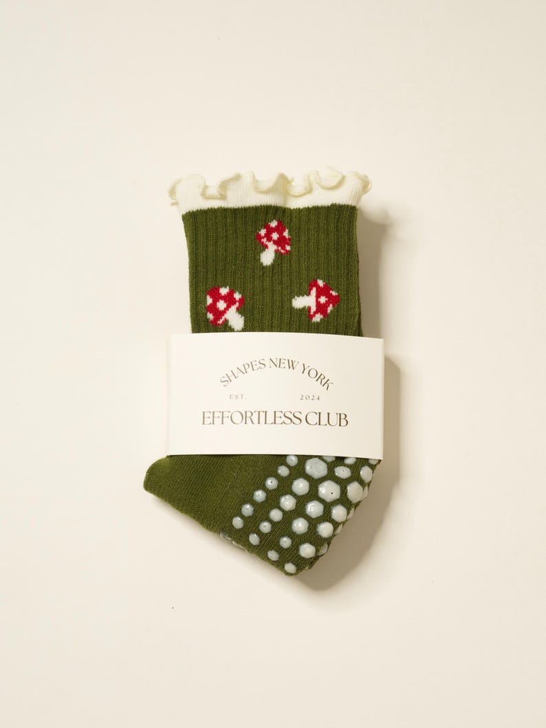 May include: Olive green socks with a white ruffled cuff and a pattern of red and white mushrooms. The socks have white non-slip grips on the sole and a white label that reads "SHAPES NEW YORK EST. 2024 EFFORTLESS CLUB."