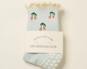 May include: Light blue socks with a white ruffle at the top and a cherry print. The socks have white grippers on the bottom and a label that reads "Shapes New York Est. 2024 Effortless Club".