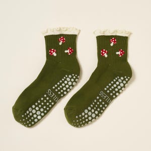 May include: A pair of olive green socks with white non-slip dots on the soles. Each sock features a white ruffled cuff and a pattern of red and white mushroom designs. The word "RITLESS" is printed on the side of each sock.
