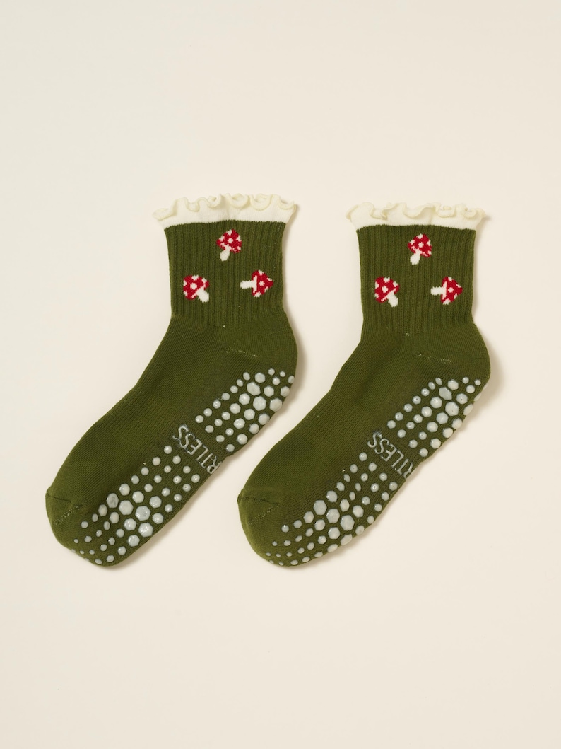 May include: A pair of olive green socks with white non-slip dots on the soles. Each sock features a white ruffled cuff and a pattern of red and white mushroom designs. The word "RITLESS" is printed on the side of each sock.