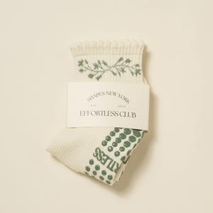 May include: Cream-coloured socks with green floral design and green dotted pattern on the sole. The socks are folded and have a tag that reads "SHAPES NEW YORK EST. 2023 EFFORTLESS CLUB."