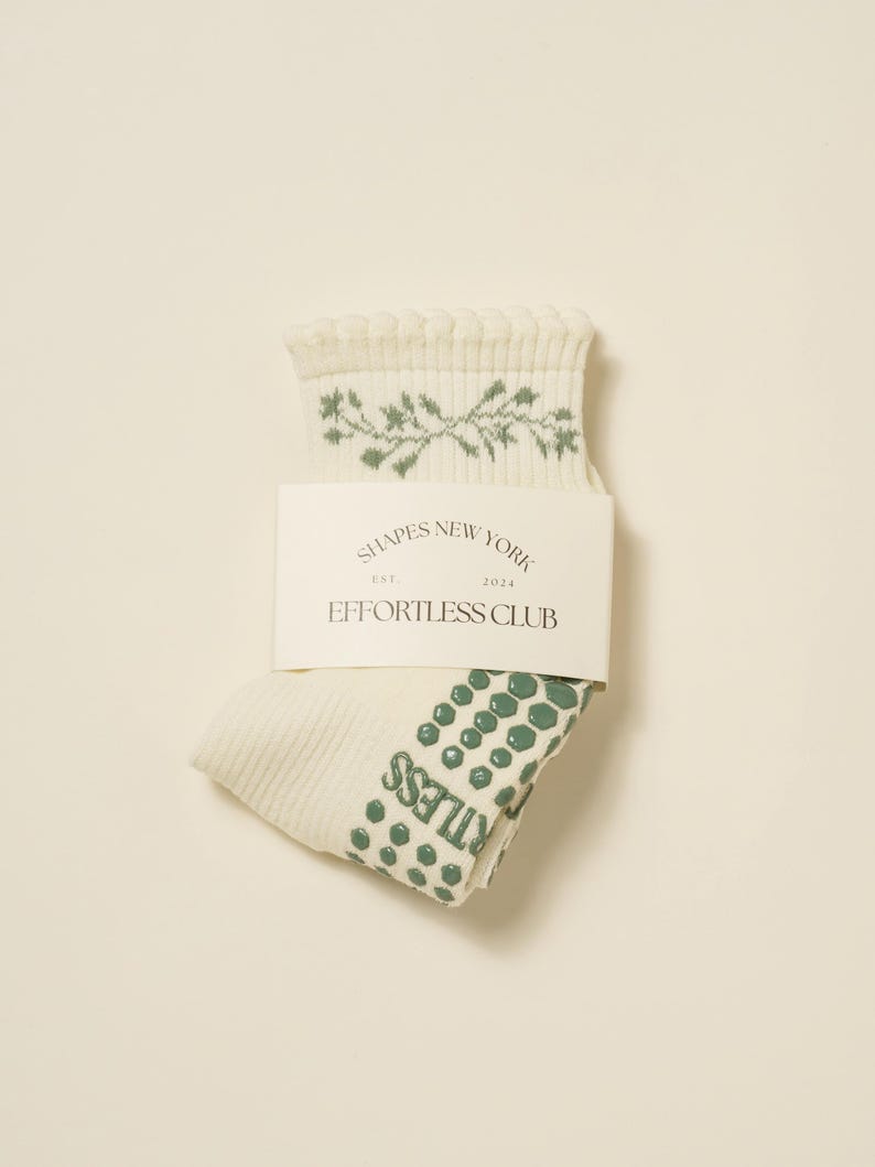 May include: Cream-coloured socks with green floral design and green dotted pattern on the sole. The socks are folded and have a tag that reads "SHAPES NEW YORK EST. 2023 EFFORTLESS CLUB."