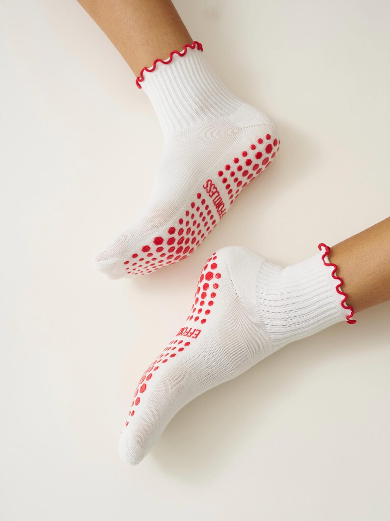 May include: White socks with red wavy trim and red dotted grips on the soles. The word "EFFORTLESS" is printed in red on the sole of each sock. The socks are ribbed and appear to be made of a soft material.