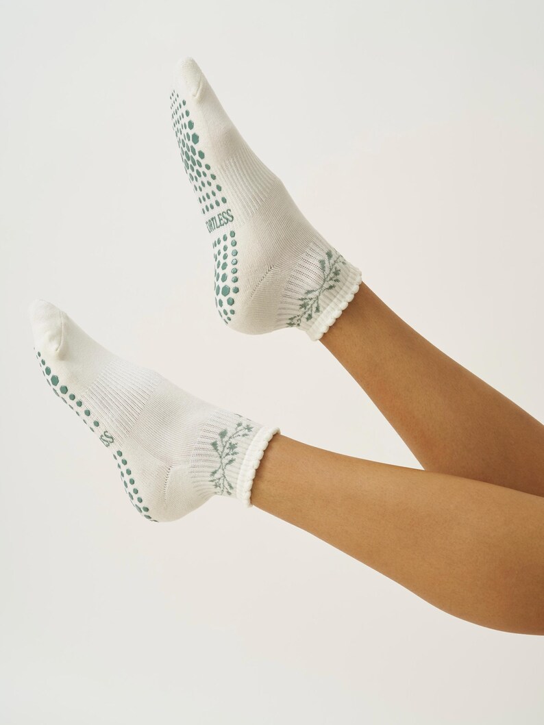 May include: White socks with green non-slip dots on the soles and the word "ORTLESS" printed on the side. The cuffs feature a green floral design. These socks are designed for comfort and grip.
