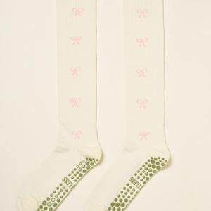May include: A pair of off-white knee-high socks with pink bow patterns. The socks have a ribbed texture and green non-slip grips on the soles. The word "LESS" is printed on the sole.