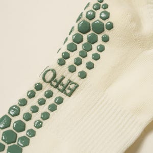 May include: Close-up of a cream-coloured sock with green hexagonal grip pads on the sole. The word "EFFO" is printed in green on the side of the sock. The sock is made of a soft, ribbed material.
