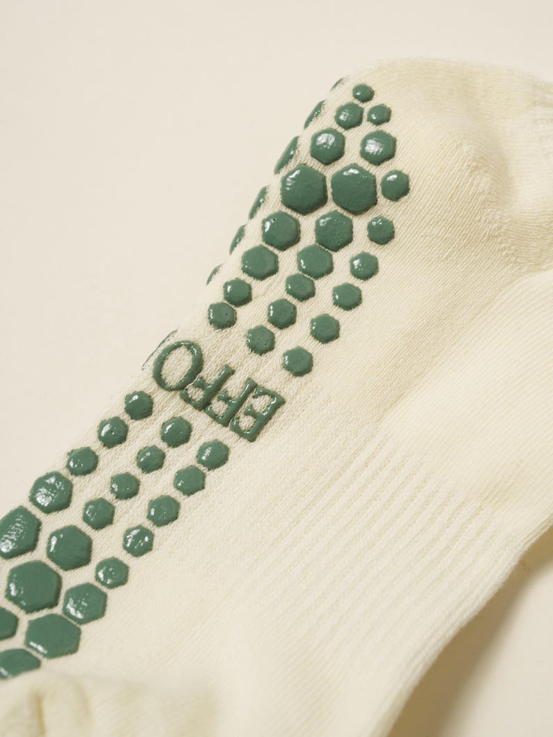May include: Close-up of a cream-coloured sock with green hexagonal grip pads on the sole. The word "EFFO" is printed in green on the side of the sock. The sock is made of a soft, ribbed material.