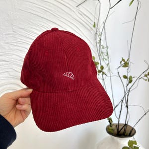 Op de afbeelding: Een rode corduroy baseballpet met een witte geborduurde halve maan op de voorkant.