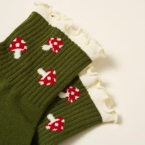 May include: A pair of green socks with a ribbed texture, featuring a pattern of red and white mushroom designs. The socks have a white ruffled trim at the top.