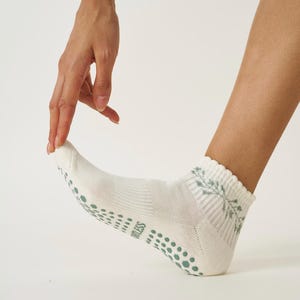 May include: White ankle socks featuring a muted green floral pattern and a ribbed cuff. The soles have a dotted grip pattern. The word "ORTLESS" is printed on the side of the sock. These socks are designed for comfort.