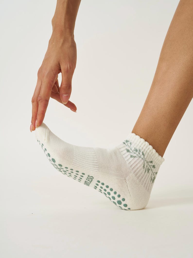 May include: White ankle socks featuring a muted green floral pattern and a ribbed cuff. The soles have a dotted grip pattern. The word "ORTLESS" is printed on the side of the sock. These socks are designed for comfort.