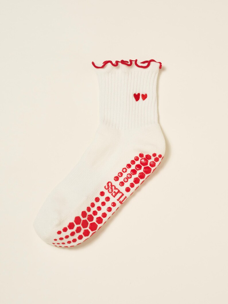 May include: White ankle socks with red accents. The socks feature a red ruffled trim at the top and two embroidered red hearts. The soles have a red, honeycomb-patterned grip with the word "TILESS" printed on the side.