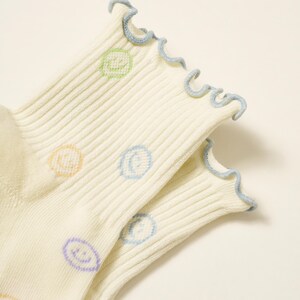 May include: A pair of off-white socks with a ribbed texture. The socks feature a light blue ruffled trim and are decorated with colorful smiley face designs in pastel shades of green, orange, blue, and purple. The socks are made of a soft, comfortable material.