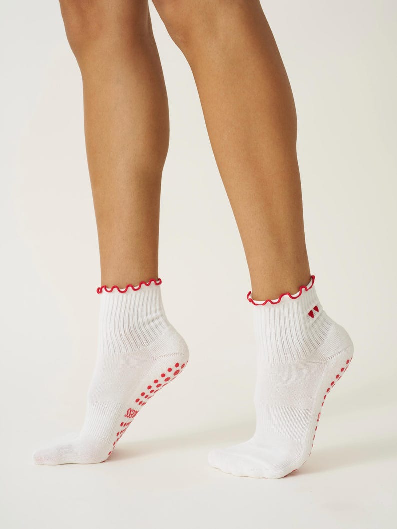 May include: White ankle socks with a red scalloped trim and red non-slip grips on the soles. The socks have a ribbed design and a small red heart detail near the ankle.