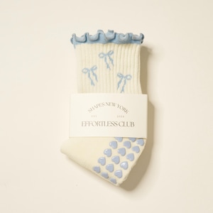 May include: A pair of cream-colored socks with light blue accents. The top edge has a ruffled trim, and the socks feature light blue bow designs. The sole has a pattern of light blue heart-shaped grips. The label reads "SHAPES NEW YORK EST. 2024 EFFORTLESS CLUB."