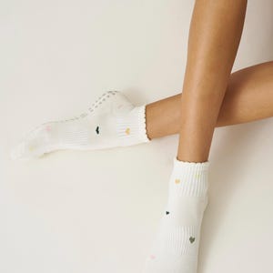 May include: White socks with small, colorful heart patterns. The socks have a ribbed design and a scalloped edge at the top. The soles have small, non-slip grips. The socks are on a light background.