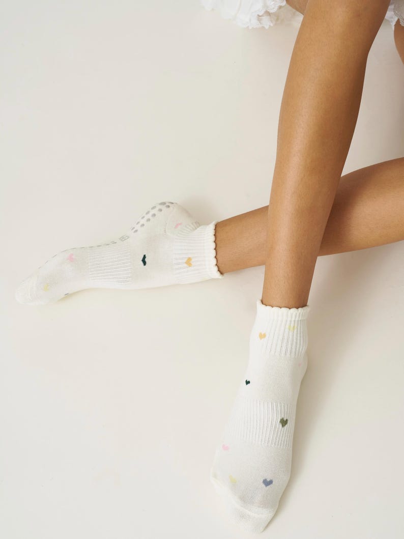 May include: White socks with small, colorful heart patterns. The socks have a ribbed design and a scalloped edge at the top. The soles have small, non-slip grips. The socks are on a light background.