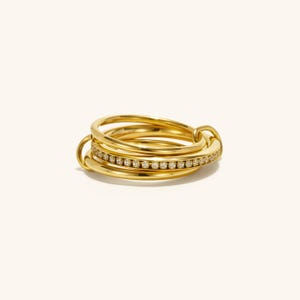 May include: A stack of gold rings with a band of small, clear stones. The rings are intertwined, creating a layered effect. The jewelry is set against a plain, light background, highlighting the gold's shine.