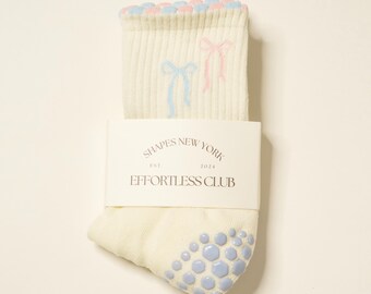 May include: Cream-colored socks with light blue and pink bow accents and a light blue grip pattern on the sole. The socks have a ribbed texture and a scalloped edge. The packaging reads "SHAPES NEW YORK EST. 2024 EFFORTLESS CLUB."