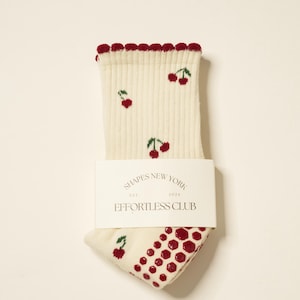 Peut inclure: Chaussettes cr&egrave;me avec un motif de cerises et une bordure festonn&eacute;e rouge. Les chaussettes ont une bande blanche avec le texte "SHAPES NEW YORK EST. 2024 EFFORTLESS CLUB". Le talon et l'orteil pr&eacute;sentent un motif &agrave; pois rouges.