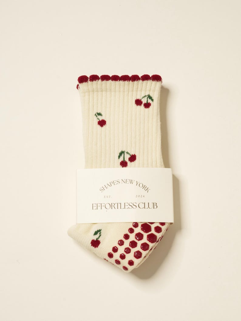 Peut inclure: Chaussettes cr&egrave;me avec un motif de cerises et une bordure festonn&eacute;e rouge. Les chaussettes ont une bande blanche avec le texte "SHAPES NEW YORK EST. 2024 EFFORTLESS CLUB". Le talon et l'orteil pr&eacute;sentent un motif &agrave; pois rouges.