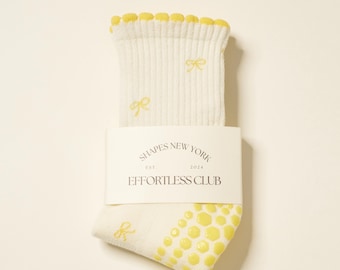 May include: Cream-colored socks with yellow bow accents and a yellow honeycomb pattern on the sole. The top edge has a yellow scalloped trim. A white label reads "SHAPES NEW YORK EST. 2024 EFFORTLESS CLUB."