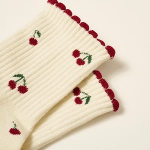 Peut inclure: Une paire de chaussettes c&ocirc;tel&eacute;es cr&egrave;me avec un motif r&eacute;p&eacute;t&eacute; de cerises rouges avec des feuilles vertes. Le bord sup&eacute;rieur des chaussettes est festonn&eacute; et bord&eacute; d'un rouge fonc&eacute;.
