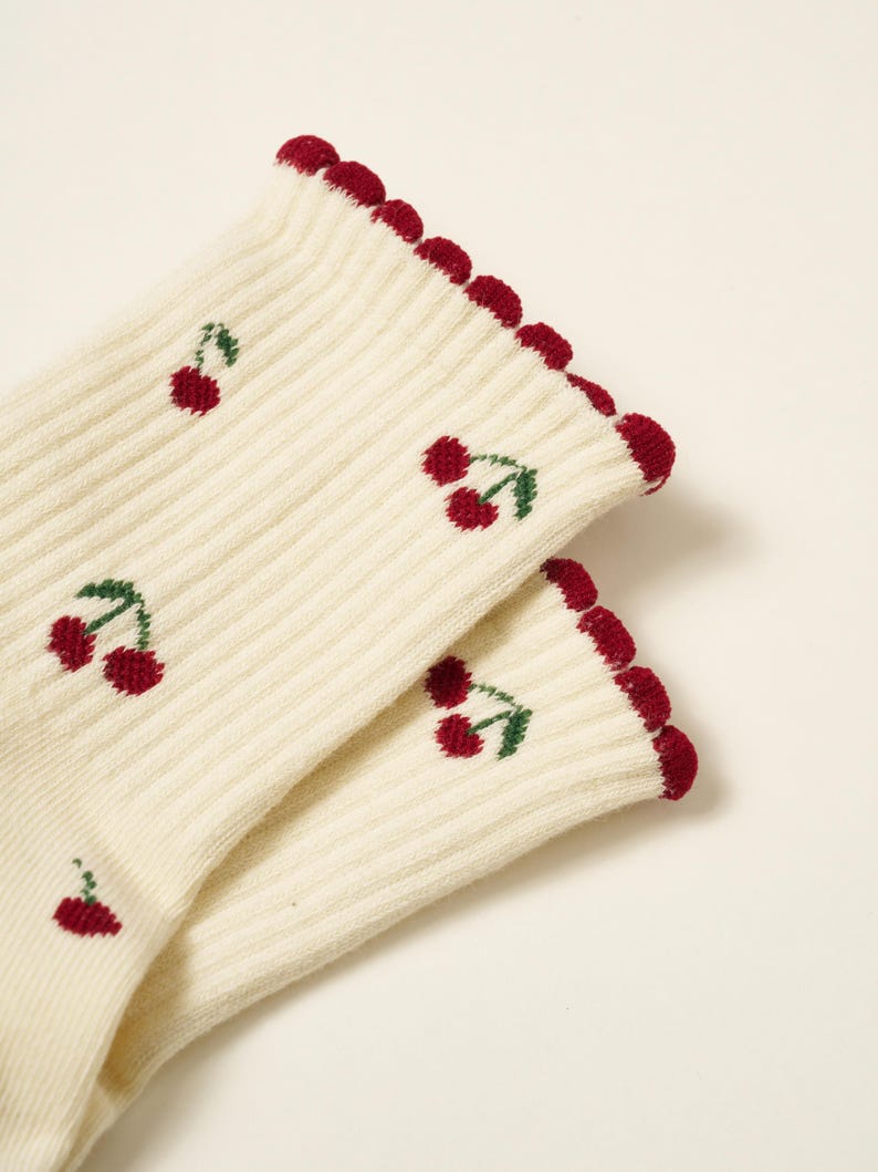 Peut inclure: Une paire de chaussettes c&ocirc;tel&eacute;es cr&egrave;me avec un motif r&eacute;p&eacute;t&eacute; de cerises rouges avec des feuilles vertes. Le bord sup&eacute;rieur des chaussettes est festonn&eacute; et bord&eacute; d'un rouge fonc&eacute;.