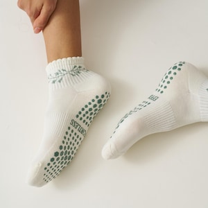 May include: White ankle socks with green non-slip dots on the soles and heels. The socks have a decorative floral pattern at the cuff and the word "FORTLESS" printed on the side. The socks are designed for comfort and grip.