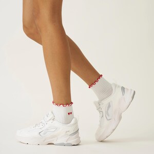 May include: White athletic shoes with a chunky sole and silver accents. The shoes are paired with white ankle socks featuring a red ruffled trim and two small red hearts. The background is a solid white.