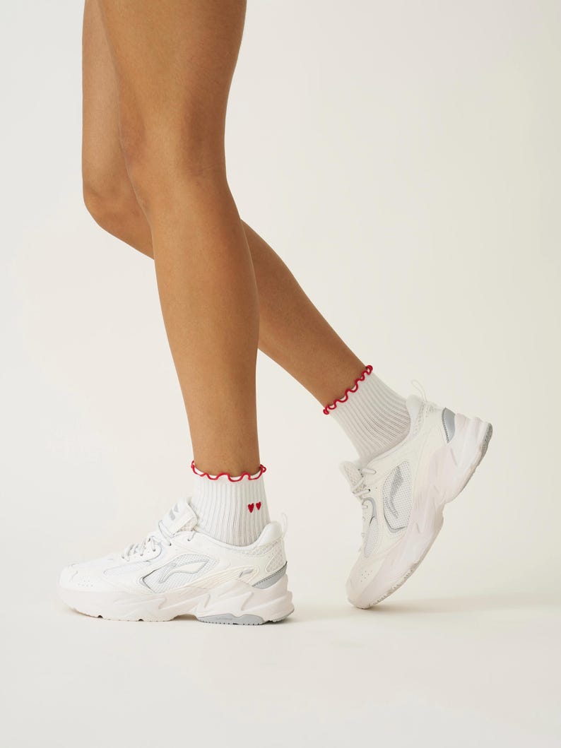 May include: White athletic shoes with a chunky sole and silver accents. The shoes are paired with white ankle socks featuring a red ruffled trim and two small red hearts. The background is a solid white.