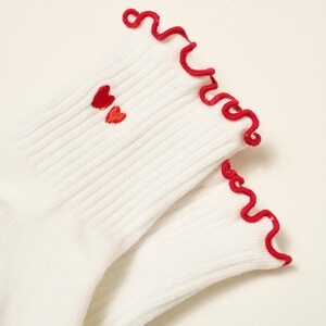 May include: A pair of white ribbed socks with red ruffled trim and two small embroidered red hearts near the ankle. The socks are made of a soft, comfortable material.