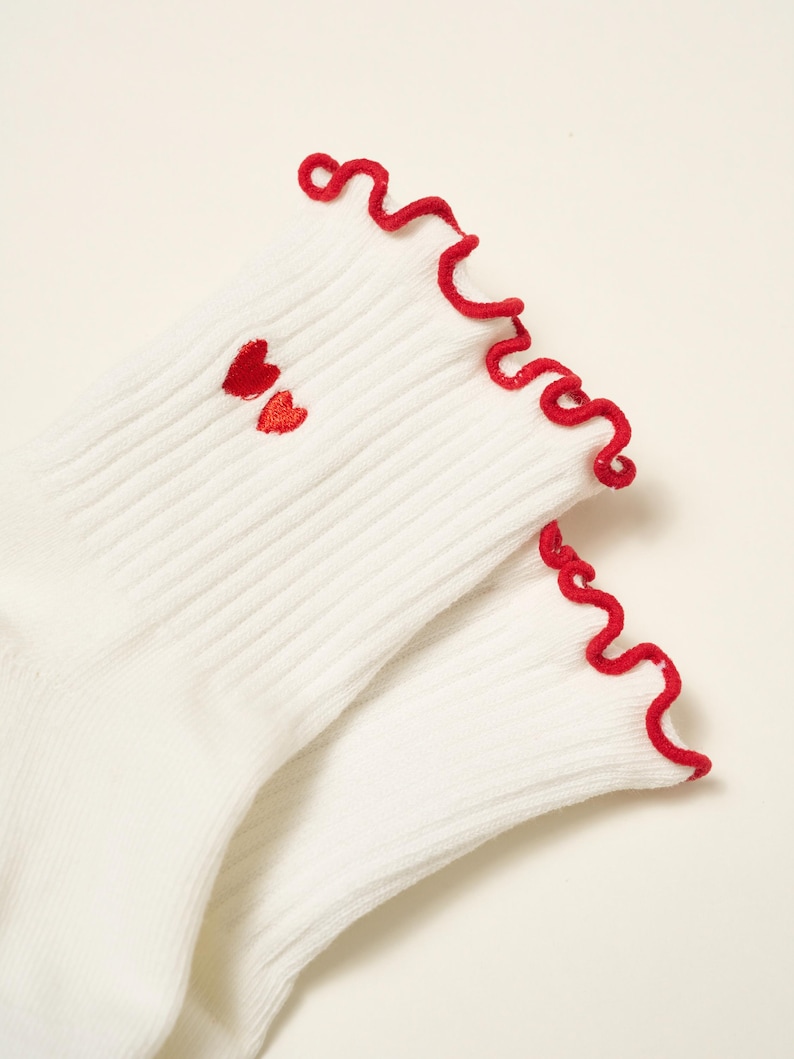 May include: A pair of white ribbed socks with red ruffled trim and two small embroidered red hearts near the ankle. The socks are made of a soft, comfortable material.