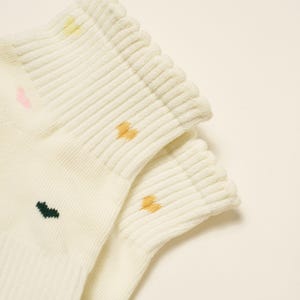 May include: Close-up of two pairs of cream-colored socks. Each sock has a ribbed cuff and small, colorful embroidered details: pink, yellow, dark green, and orange. The socks are made of a soft, textured material.