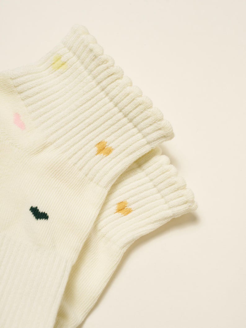 May include: Close-up of two pairs of cream-colored socks. Each sock has a ribbed cuff and small, colorful embroidered details: pink, yellow, dark green, and orange. The socks are made of a soft, textured material.