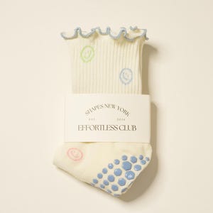 Puede incluir: Calcetines color crema con textura acanalada, con diseños de caras sonrientes en verde, azul y rosa. El borde superior tiene un ribete con volantes y un borde azul claro. El embalaje incluye el texto "SHAPES NEW YORK EST. 2024 EFFORTLESS CLUB."