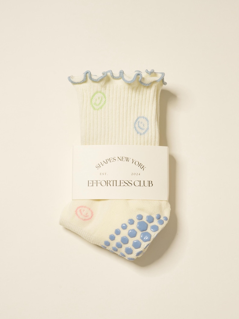 May include: Cream-colored socks with a ribbed texture, featuring smiley face designs in green, blue, and pink. The top edge has a ruffled trim with a light blue border. The packaging includes the text "SHAPES NEW YORK EST. 2024 EFFORTLESS CLUB."