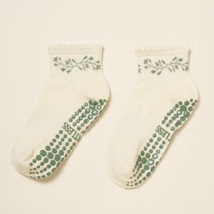 May include: Two off-white ankle socks with a sage green floral pattern at the cuff. The soles have a pattern of green hexagonal grips and the word "LESS". These socks are designed for comfort and grip.