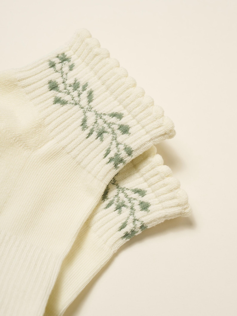 May include: A pair of cream-coloured socks with a ribbed cuff. The cuff features a delicate green floral pattern. The socks are made of a soft, textured material, and the design is simple and elegant.