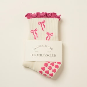May include: White socks with pink bows and hearts. The socks have a frilly pink trim at the top. The socks are labeled "Shapes New York Est. 2024 Effortless Club".