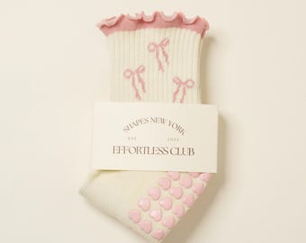 May include: White socks with pink bows and pink heart-shaped grips on the bottom. The socks have a pink ruffled trim at the top. The socks are packaged with a white label that reads "Shapes New York Est. 2024 Effortless Club".
