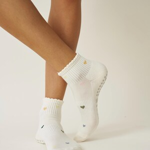 May include: White ankle socks with a scalloped edge and small, colorful heart patterns. The socks have a ribbed cuff and a non-slip grip on the sole. The hearts are in shades of green, yellow, and pink.