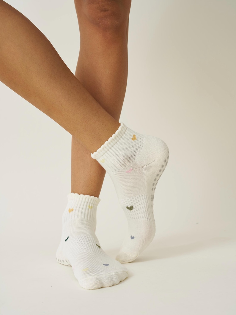 May include: White ankle socks with a scalloped edge and small, colorful heart patterns. The socks have a ribbed cuff and a non-slip grip on the sole. The hearts are in shades of green, yellow, and pink.