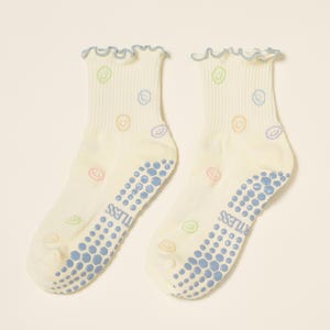 May include: A pair of off-white socks with a light blue ruffled trim and a pattern of colorful smiley faces. The soles have blue non-slip grips with the word "TUTLESS" printed on them.