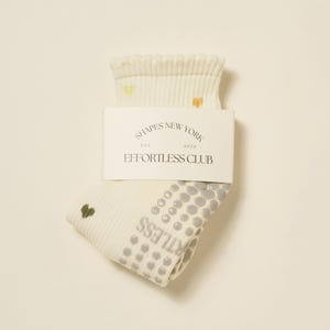 May include: Cream-colored socks with a ribbed cuff and small heart accents. The socks have a non-slip grip pattern on the sole and the text "EFFORTLESS CLUB" printed on the side. A white label with the text "SHAPES NEW YORK" is attached.