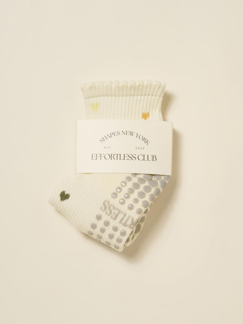 May include: Cream-colored socks with a ribbed cuff and small heart accents. The socks have a non-slip grip pattern on the sole and the text "EFFORTLESS CLUB" printed on the side. A white label with the text "SHAPES NEW YORK" is attached.